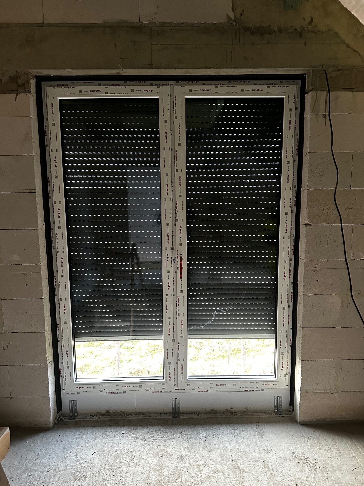Białe okno PCV Aluplast z opuszczoną roletą zewnętrzną w surowym murze z betonu komórkowego. Widoczne mocowania okna. Częściowo widoczny widok za oknem.