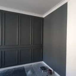 Świeżo pomalowane na ciemny szary kolor panele ścienne z dekoracyjnymi listwami, widoczne gniazdka elektryczne i folia ochronna na podłodze, obok puszka farby i kuweta malarska.