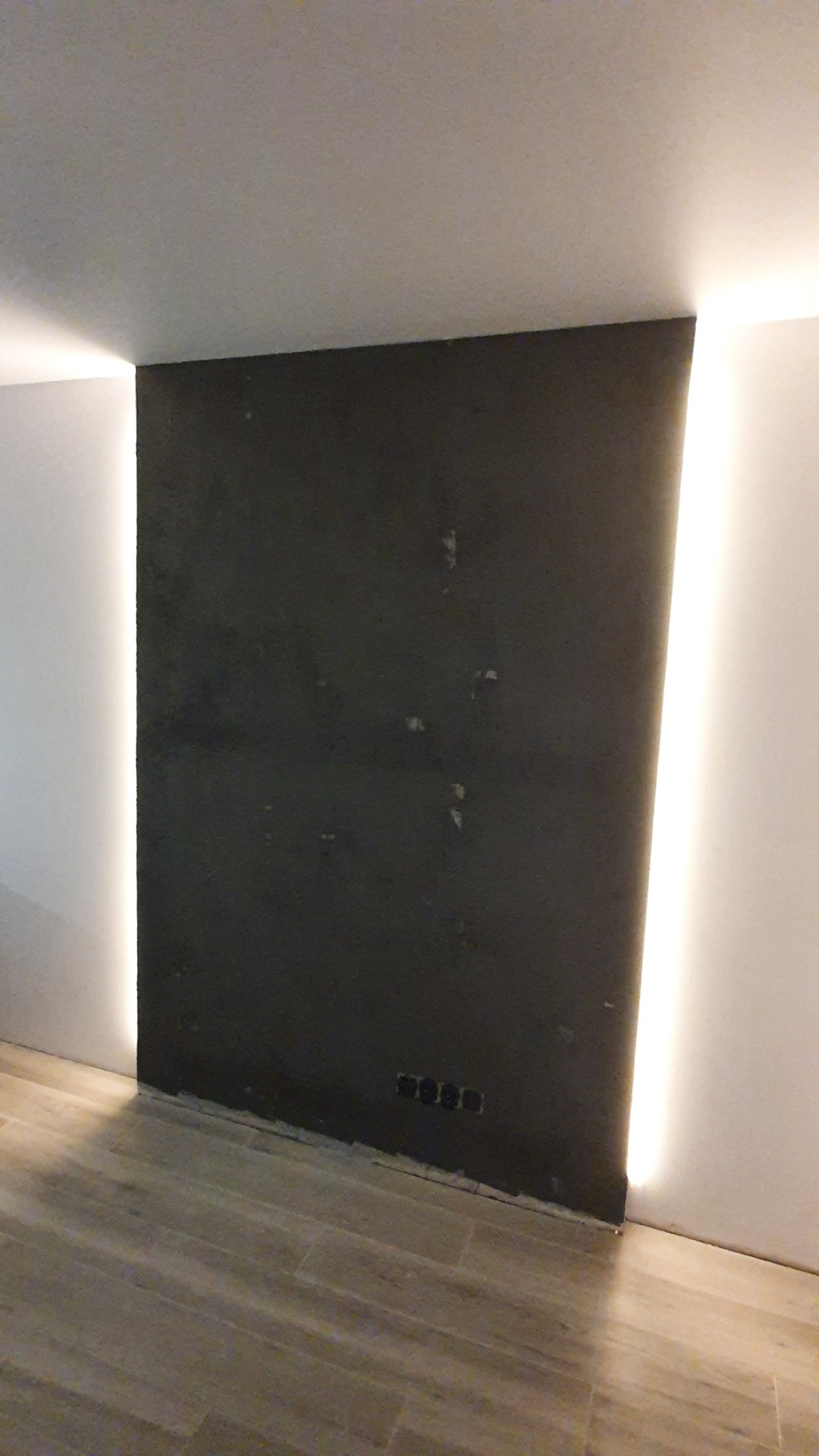 Ciemna ściana z surowym wykończeniem i wbudowanym oświetleniem LED po bokach, widoczne gniazdka elektryczne przy podłodze, na podłodze płytki imitujące drewno.