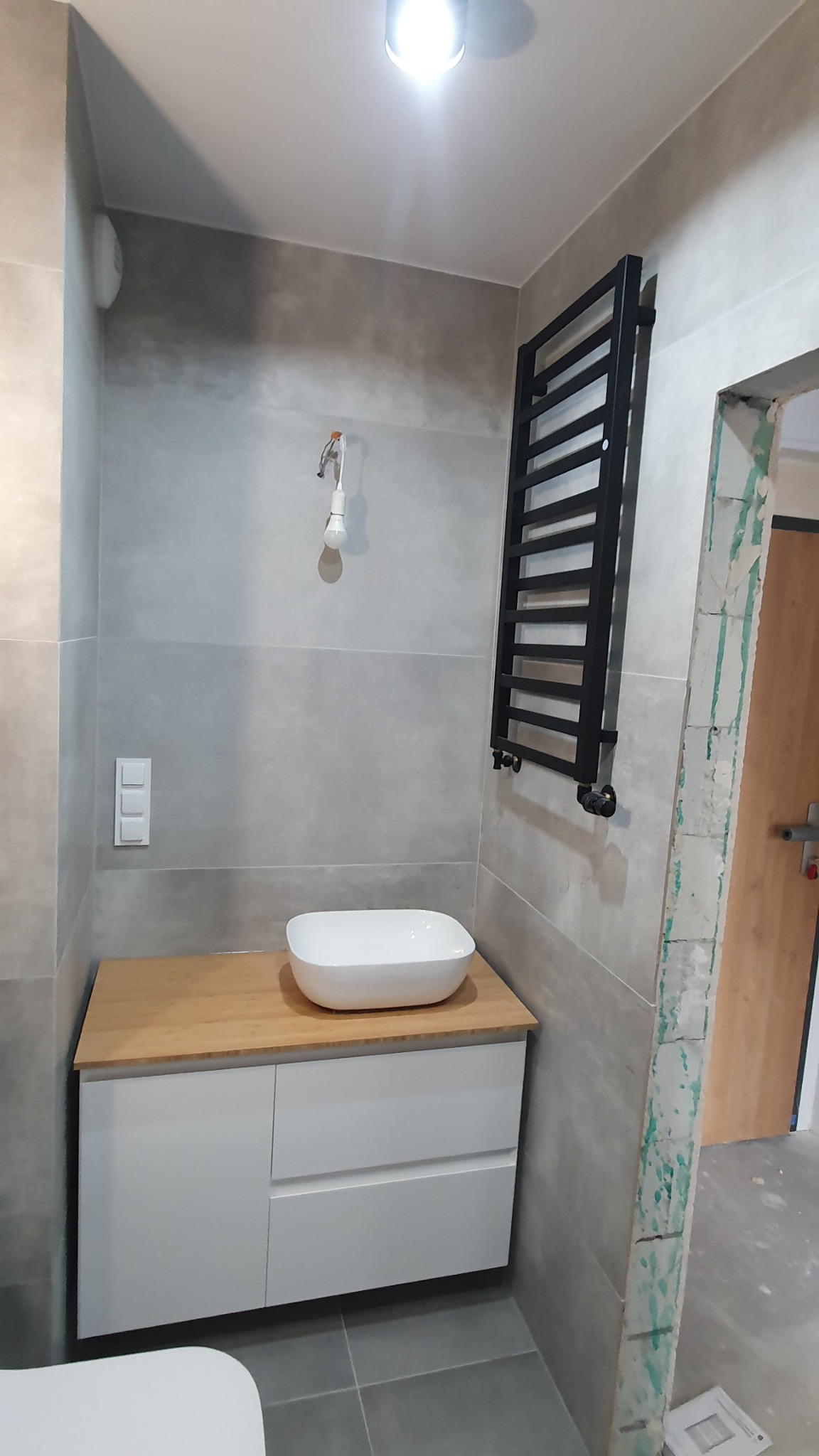 Łazienka w trakcie wykańczania: szare płytki, biała szafka z drewnianym blatem pod umywalkę, czarny grzejnik drabinkowy, wystająca żarówka na kablu, widoczne ślady po pracach budowlanych przy...