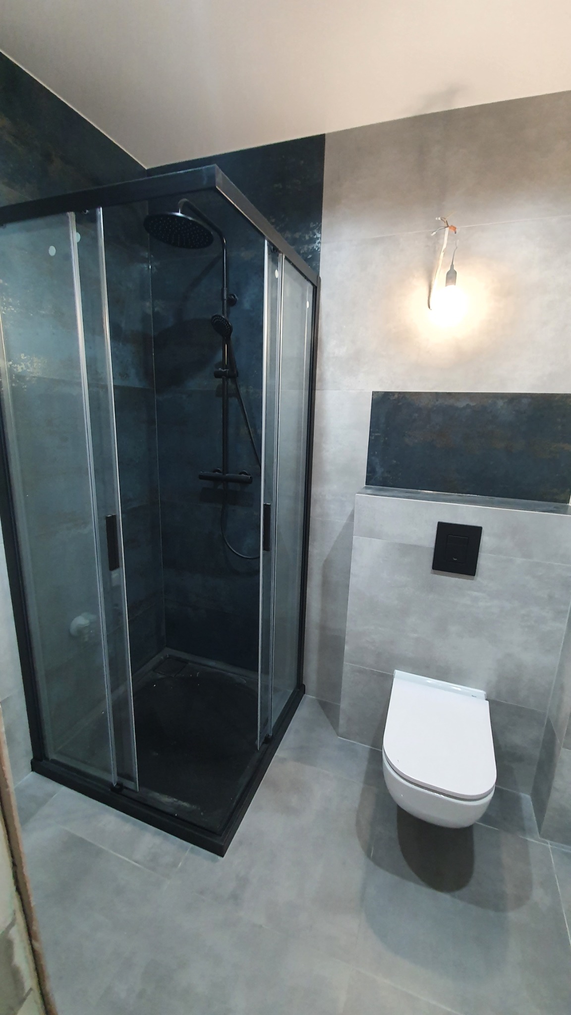 Nowoczesna kabina prysznicowa z czarną armaturą i przesuwnymi drzwiami w łazience w Kiełczowie, wykończonej szarymi płytkami, z widoczną podwieszaną toaletą i niedokończonym oświetleniem.