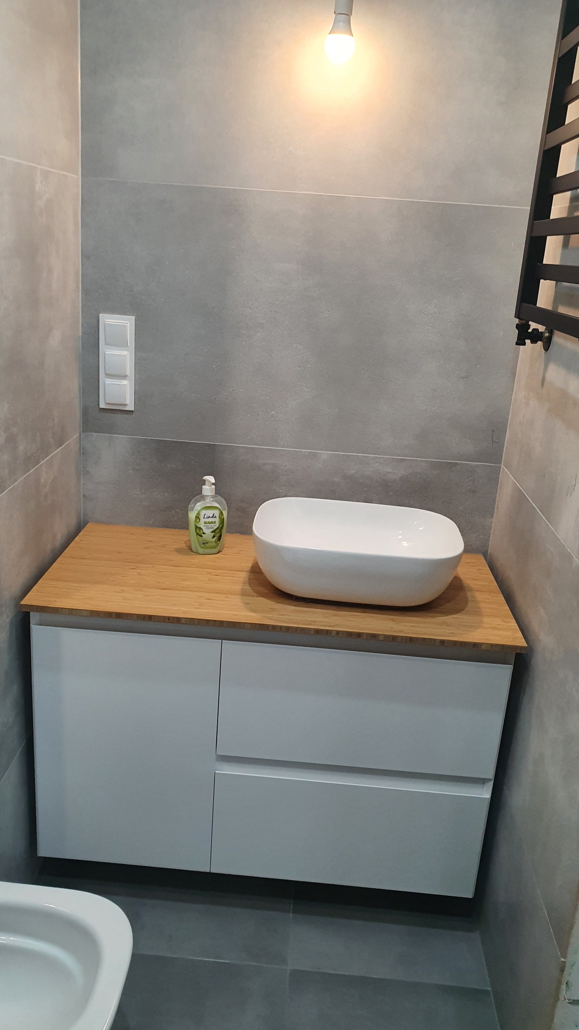 Umywalka nablatowa w kształcie łódki na drewnianym blacie szafki łazienkowej z szufladami, szare płytki ścienne i podłogowe, butelka mydła w płynie.