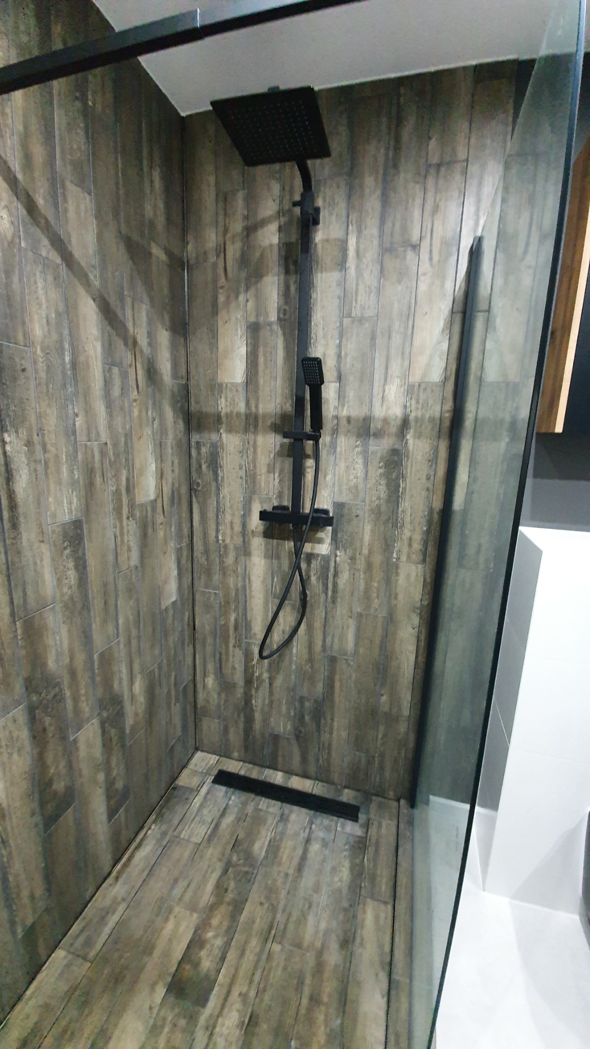 Kabina prysznicowa typu walk-in z czarną armaturą i odpływem liniowym, wykończona płytkami imitującymi drewno.