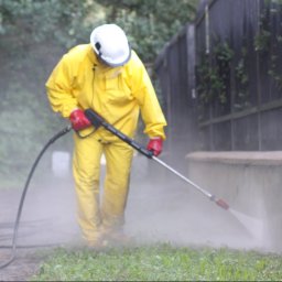 500BAR SPÓŁKA Z O.O. - Pracownik w żółtym kombinezonie, kasku i czerwonych rękawicach czyści elewację niskiego murku za pomocą myjki ciśnieniowej, w tle widoczny zielony trawnik i ciemne ogrodzenie.