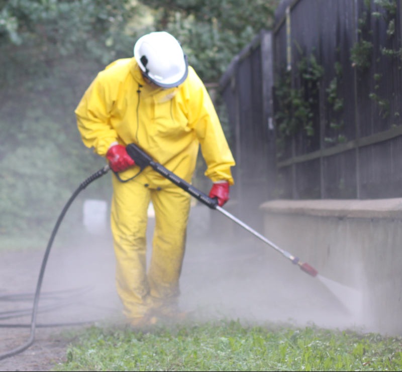 Pracownik w żółtym kombinezonie, kasku i czerwonych rękawicach czyści elewację niskiego murku za pomocą myjki ciśnieniowej, w tle widoczny zielony trawnik i ciemne ogrodzenie.
