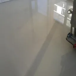 Świeżo położone, beżowe płytki podłogowe z widocznymi fugami, odbijające światło wpadające przez duże okno balkonowe, z fragmentem białej ściany z cegły dekoracyjnej i urządzenia do polerowania...