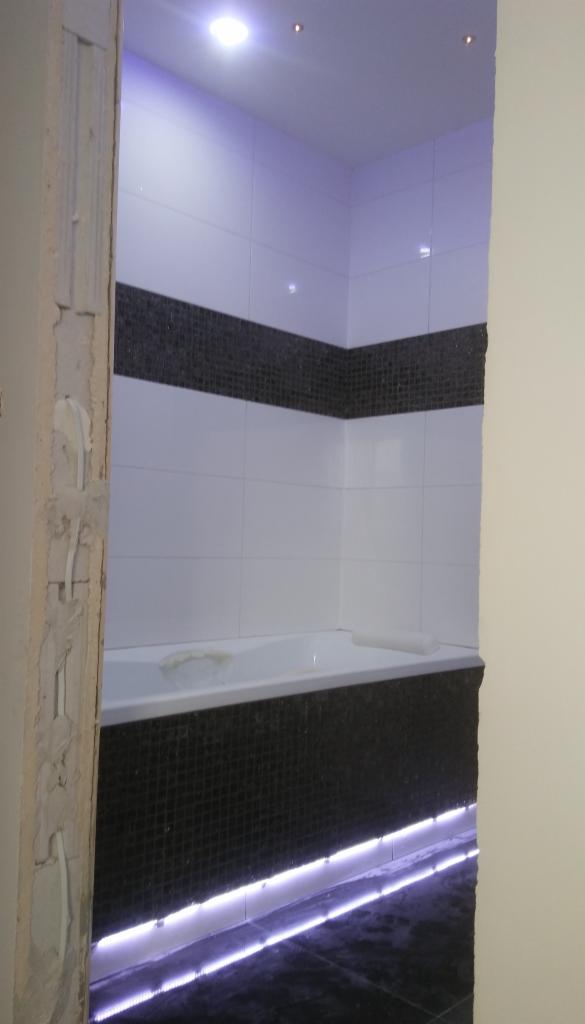 Wykończona łazienka z biało-czarnymi płytkami, podświetleniem LED pod wanną i niedokończonym ościeżem drzwi po lewej stronie.