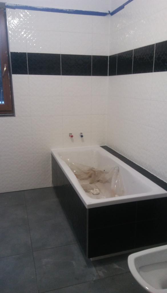 Łazienka w trakcie remontu: białe płytki z poziomym pasem czarnych płytek, zabudowana wanna z folią ochronną w środku, szare płytki podłogowe, widoczne wystające rury do baterii.