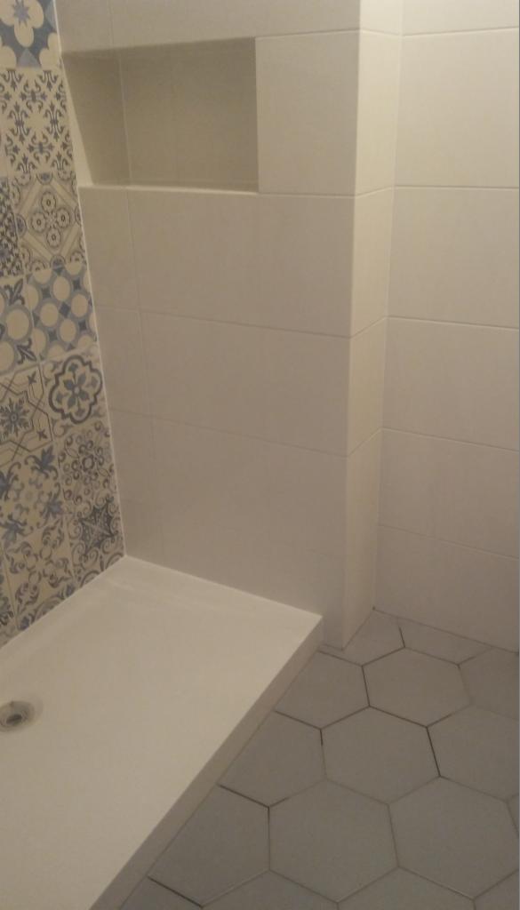 Wykończona łazienka z kabiną prysznicową: białe, prostokątne płytki na ścianach, dekoracyjne płytki z motywem kwiatowym przy kabinie, sześciokątne szare płytki podłogowe i wnęka w ścianie.