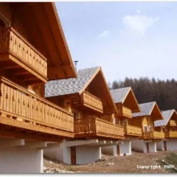 Rząd identycznych, parterowych domów z drewna z balkonami, osadzonych na betonowych fundamentach, na tle zalesionego wzgórza.