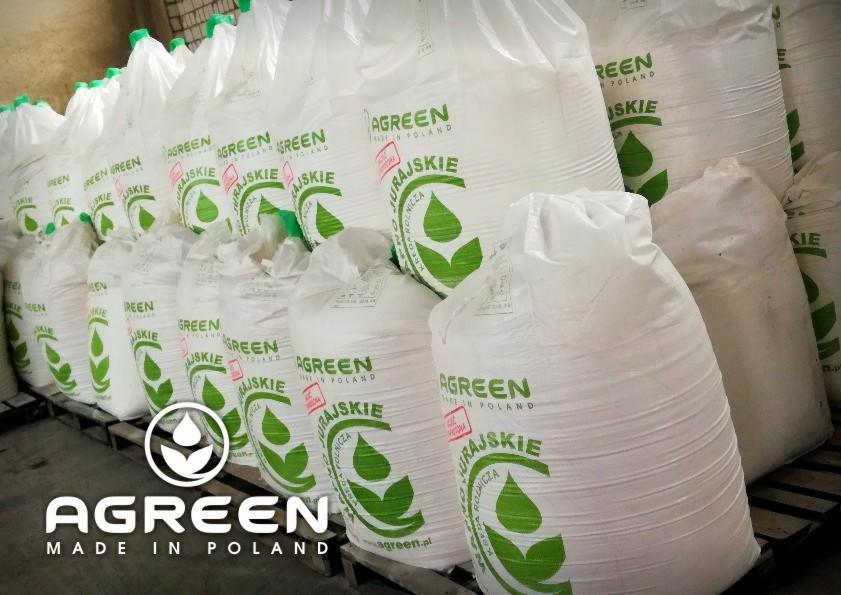 Stos białych worków z zielonym nadrukiem logo producenta, ułożonych na paletach w magazynie.