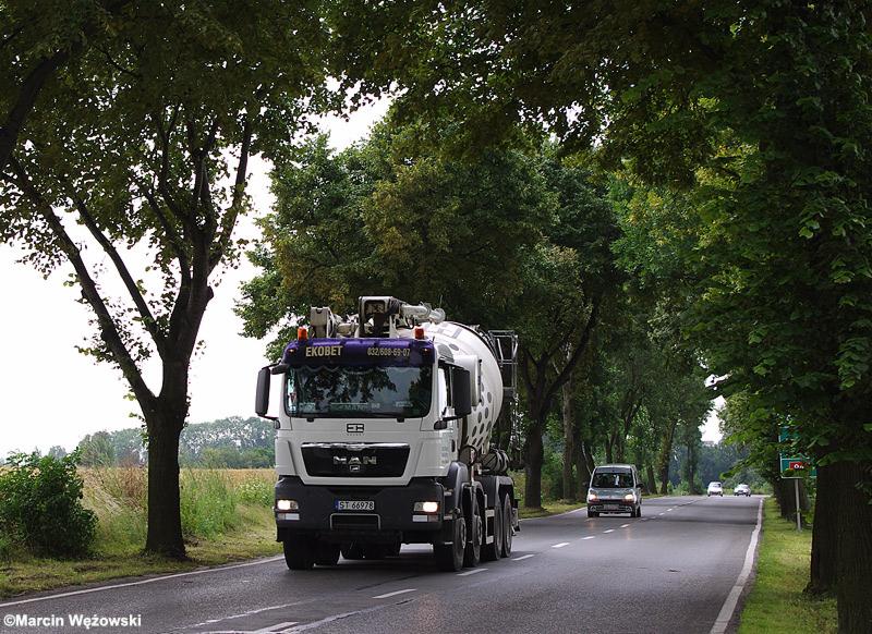 Ciężarówka z betonomieszarką marki MAN, z napisem EKOBET na kabinie, jadąca asfaltową drogą w otoczeniu drzew.
