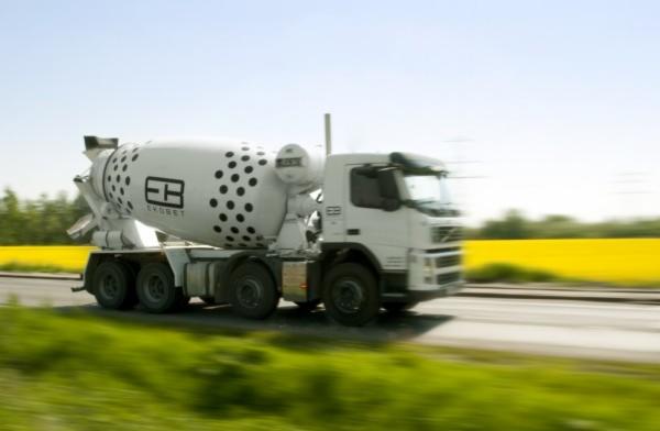 Biała betonomieszarka z czarnymi kropkami i logo firmy z Bełchatowa w ruchu, widoczna na tle żółtego pola i zieleni przydrożnej.