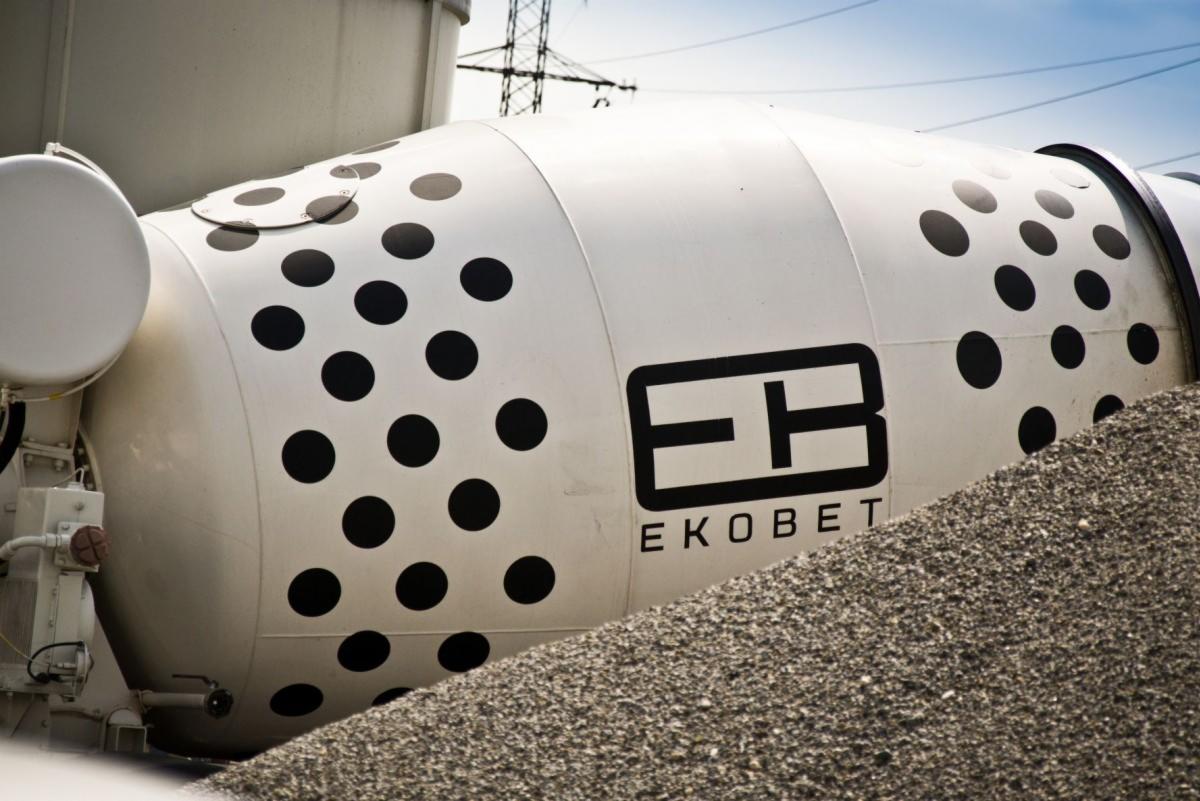 Cysterna betonomieszarki z czarnymi kropkami i logo firmy EKOBET, częściowo zasypana żwirem.