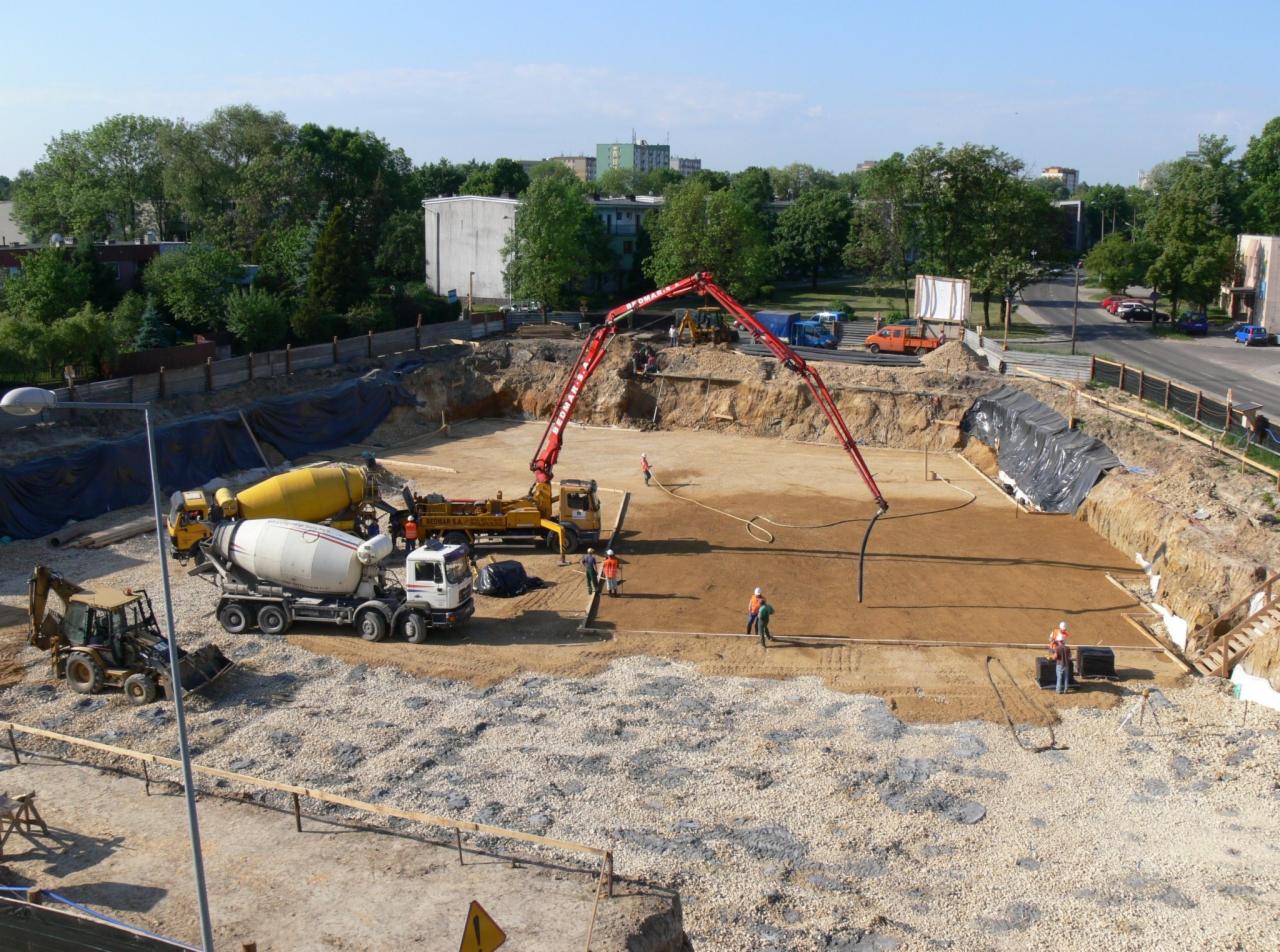 Wysoki kąt widzenia na plac budowy z pracownikami, pompą do betonu z czerwonym ramieniem, betonomieszarkami i wykopem przygotowanym pod wylewkę fundamentów.