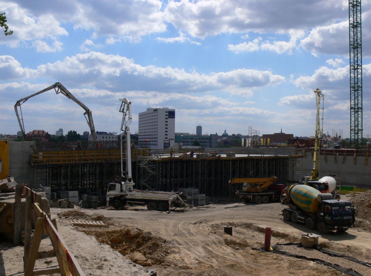 Szeroki kadr placu budowy z widocznymi: pompą do betonu na białym samochodzie, żółto-zieloną betonomieszarką, dźwigiem, zbrojeniami, deskowaniem oraz budynkami w tle pod błękitnym niebem z chmurami.