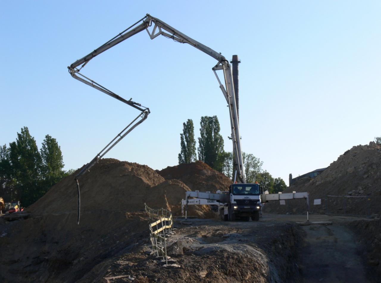 Ciężarówka z pompą do betonu na placu budowy, ramię pompy skierowane w stronę pryzmy piasku, widoczne drzewa w tle pod błękitnym niebem.