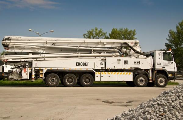 Biała ciężarówka z pompą do betonu EkoBet zaparkowana na betonowym placu, z widoczną stertą kruszywa i drzewami w tle.