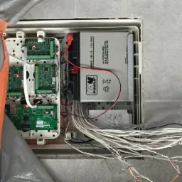 Otwarta skrzynka sterująca z elektroniką, akumulatorem 12V i plątaniną białych kabli na tle szarej ściany z płytek, częściowo zakryta pomarańczową folią ochronną.