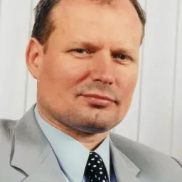 Portret biznesowy mężczyzny w szarym garniturze i krawacie w kropki, działającego w Warszawie, z delikatnym uśmiechem i poważnym wyrazem twarzy.