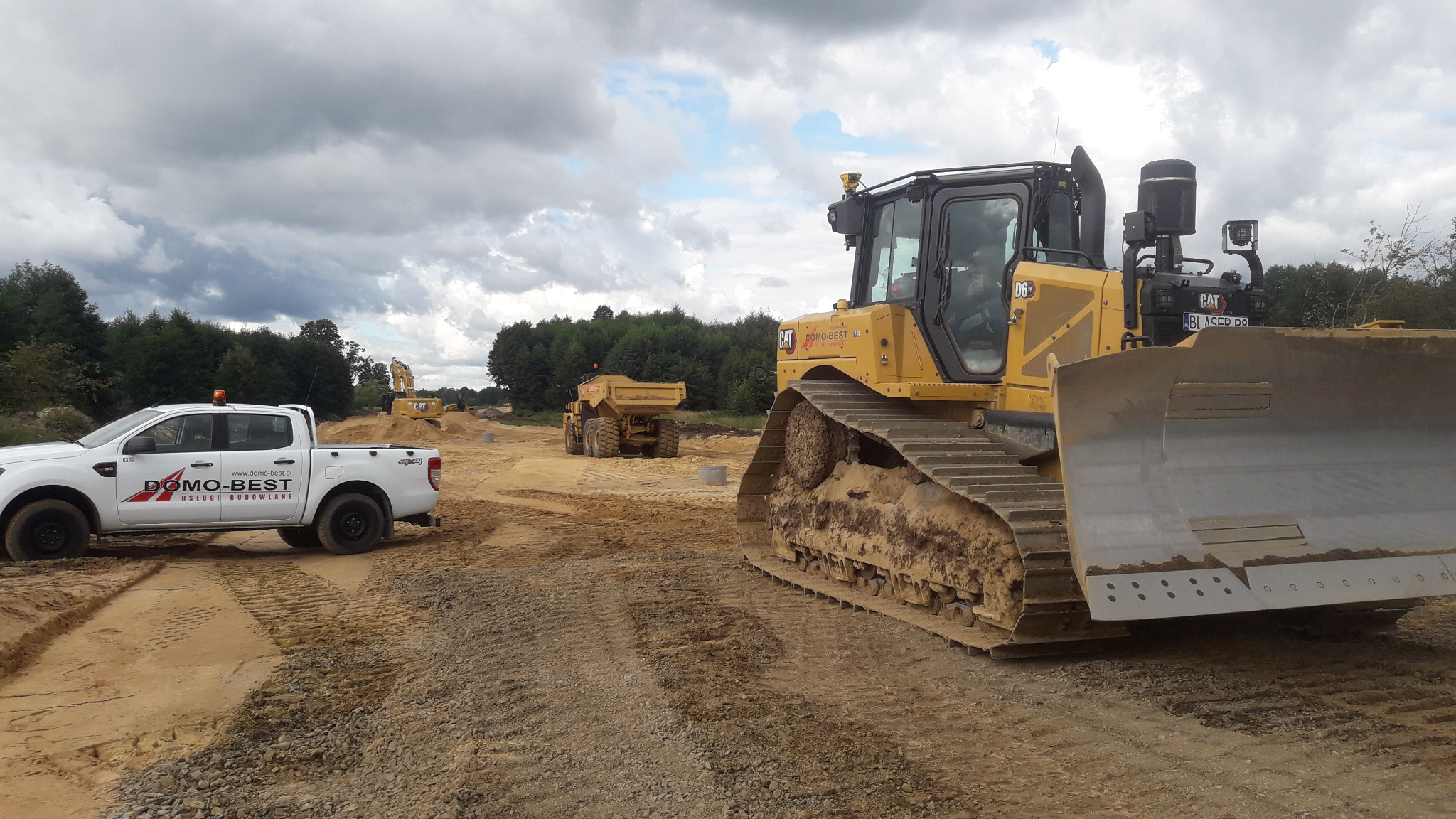 Żółty spychacz CAT na placu budowy, w tle ciężarówka i koparka. Biały pick-up z logo firmy Domo-Best z boku kadru. Pochmurne niebo nad terenem robót.