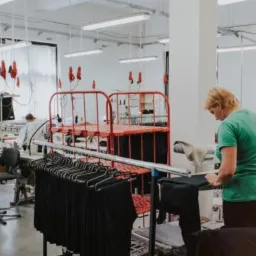 Jasne wnętrze szwalni: pracownice w zielonych koszulkach przy maszynach, w tle wieszaki z gotową odzieżą i wózki transportowe.