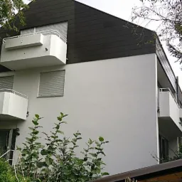 Wielopiętrowy budynek mieszkalny z białymi ścianami i ciemnym dachem, widoczne balkony z białymi barierkami i opuszczone rolety w oknach, otoczony zielenią.