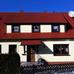 Dom jednorodzinny z jasną elewacją i czerwoną dachówką, widoczna antena satelitarna na dachu, komin i okna z firankami.