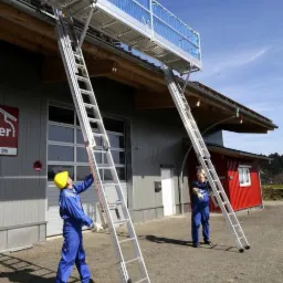Dwóch pracowników w kombinezonach ustawia aluminiową platformę roboczą z drabinkami przy budynku z szarą elewacją i czerwonym dachem.