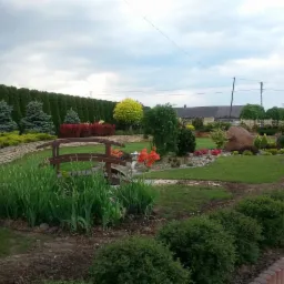 Ogród z drewnianym mostkiem nad rabatą, kamienny murek, tuje w tle, różnorodne rośliny i trawnik.