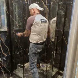 Pracownik w czapce, z tatuażami na ręce, wygładza silikon wokół kabiny prysznicowej z czarnymi płytkami imitującymi marmur, widoczne narzędzia i resztki materiałów budowlanych.