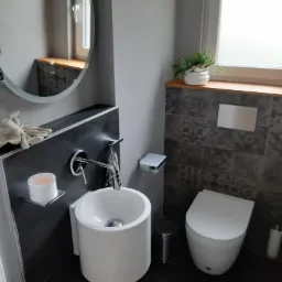 Nowoczesna, minimalistyczna łazienka w szarościach z okrągłym lustrem, wiszącą umywalką o cylindrycznym kształcie, toaletą z ukrytym spłukiwaniem i dekoracyjnymi płytkami na ścianie.