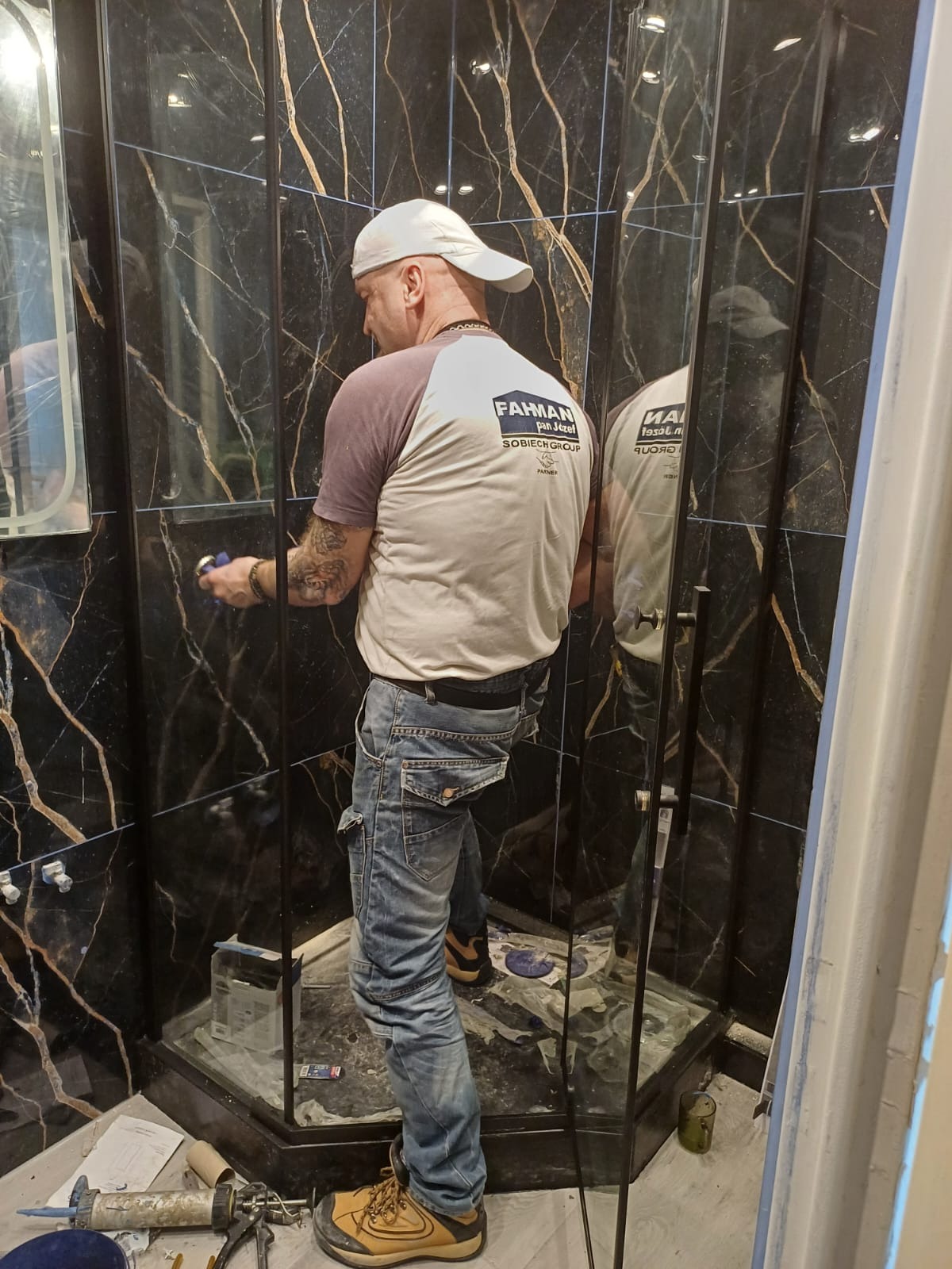 Pracownik w czapce, z tatuażami na ręce, wygładza silikon wokół kabiny prysznicowej z czarnymi płytkami imitującymi marmur, widoczne narzędzia i resztki materiałów budowlanych.