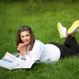 Uśmiechnięta kobieta w białej koszuli i czarnych spodniach leży na trawie, czytając gazetę, z okularami przeciwsłonecznymi na głowie i białymi tenisówkami z żółtymi podeszwami