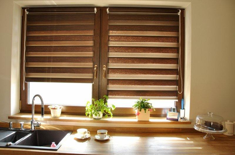 Okno z brązowymi roletami dzień noc w kuchni. Na parapecie rośliny doniczkowe, filiżanki i ciasto pod kloszem. Ujęcie z bliska, ciepłe światło.