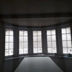 Półokrągłe okno z białymi ramami, zabezpieczone folią ochronną, w surowym wnętrzu po tynkowaniu ścian.