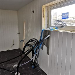 Alfa Entreprenør AS - Wnętrze w trakcie budowy, widoczne pionowe panele ścienne, okno z pianką montażową, czarna podłoga z białymi fugami, instalacja grzewcza podłogowa z rurami i folią aluminiową.