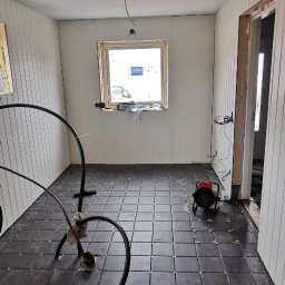 Alfa Entreprenør AS - Wnętrze w trakcie wykańczania: ciemne kwadratowe płytki na podłodze z krzyżykami dystansowymi, ściany z boazerią, okno z pianką montażową, grzejnik elektryczny i przewody instalacji podłogowej.