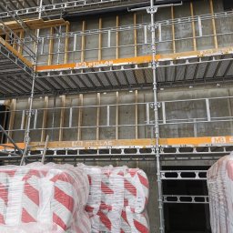 Alfa Entreprenør AS - Wnętrze budynku w trakcie budowy z widocznymi elementami konstrukcyjnymi ścian, szalunkiem i rusztowaniami, oraz paletami materiałów budowlanych zabezpieczonych folią. Widoczny etap prac...