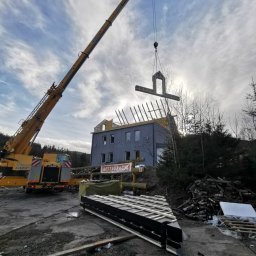 Alfa Entreprenør AS - Montaż elementu szkieletowego domu z użyciem dźwigu na tle pochmurnego nieba i częściowo wykończonego budynku.