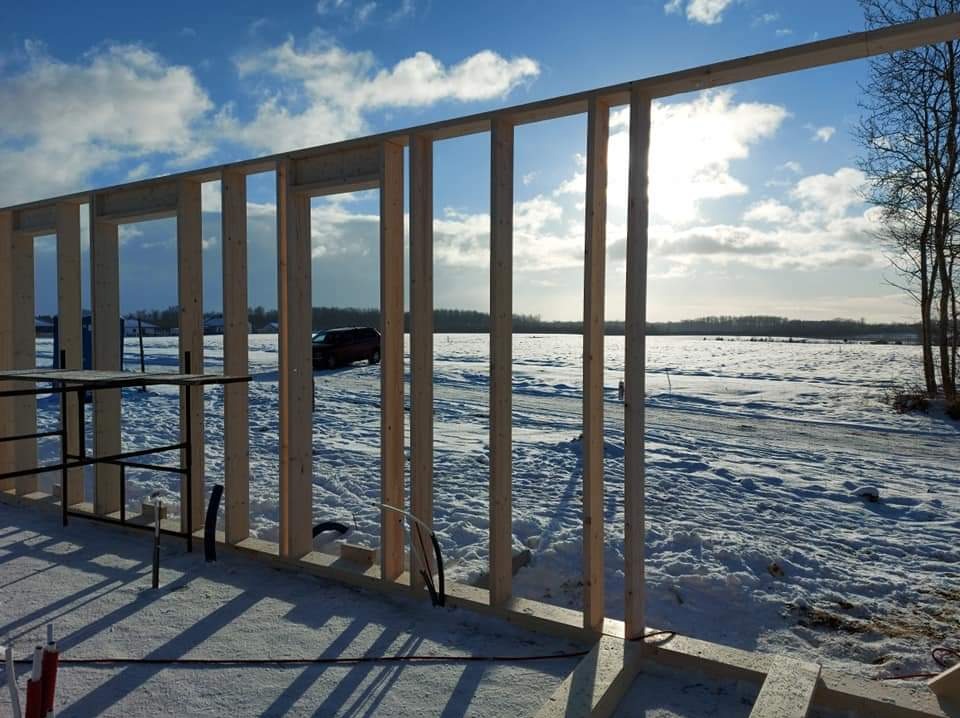 Wznoszenie drewnianej konstrukcji szkieletowej domu na zasypanym śniegiem terenie; widoczne słupy, nadproża, oraz elementy instalacji; w tle krajobraz zimowy z drzewami i samochodem.