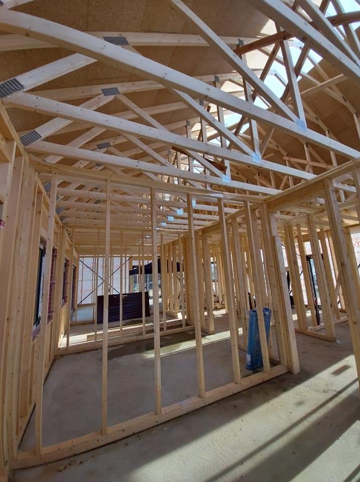 Wnętrze domu w trakcie budowy, widoczne drewniane elementy konstrukcyjne ścian i dachu połączone metalowymi złączkami, surowy beton na podłodze, niebieski zwój folii budowlanej w rogu.