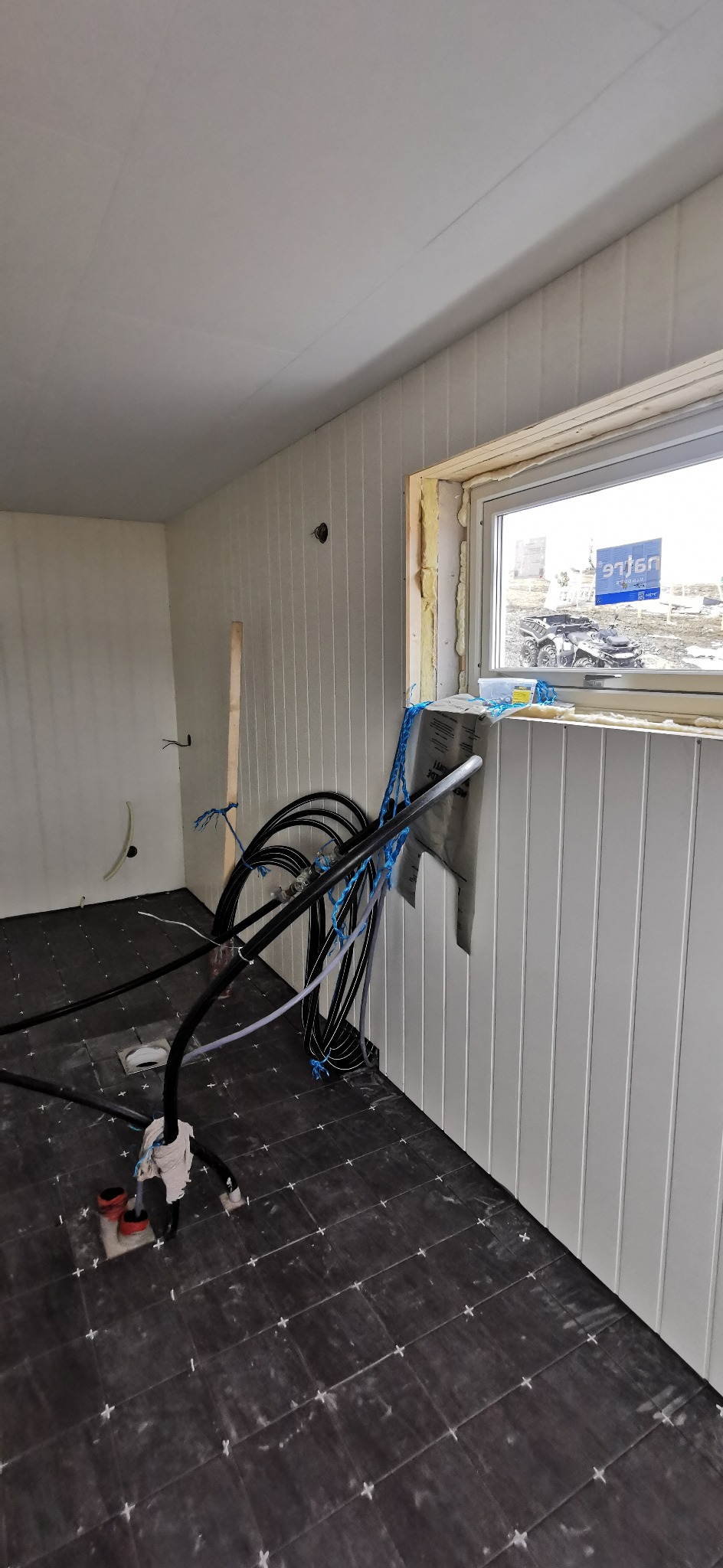 Wnętrze w trakcie budowy, widoczne pionowe panele ścienne, okno z pianką montażową, czarna podłoga z białymi fugami, instalacja grzewcza podłogowa z rurami i folią aluminiową.