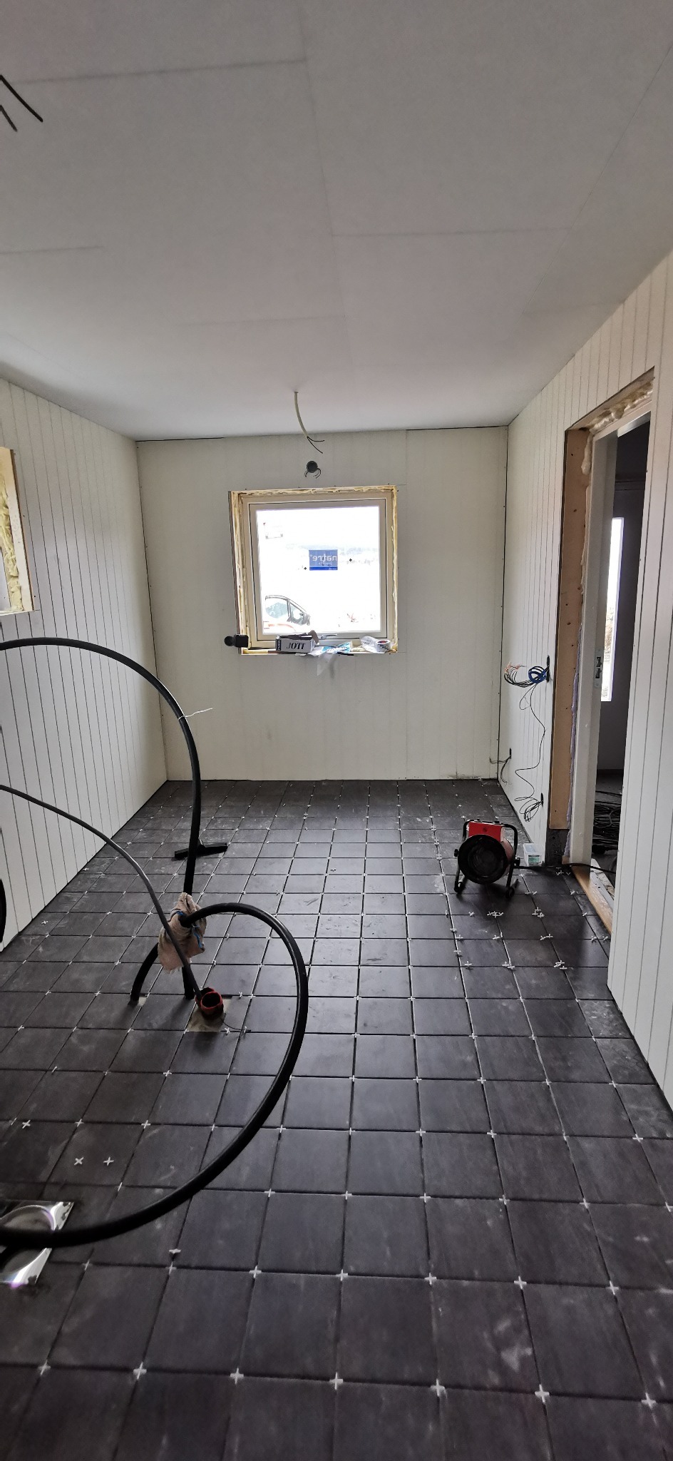 Wnętrze w trakcie wykańczania: ciemne kwadratowe płytki na podłodze z krzyżykami dystansowymi, ściany z boazerią, okno z pianką montażową, grzejnik elektryczny i przewody instalacji podłogowej.