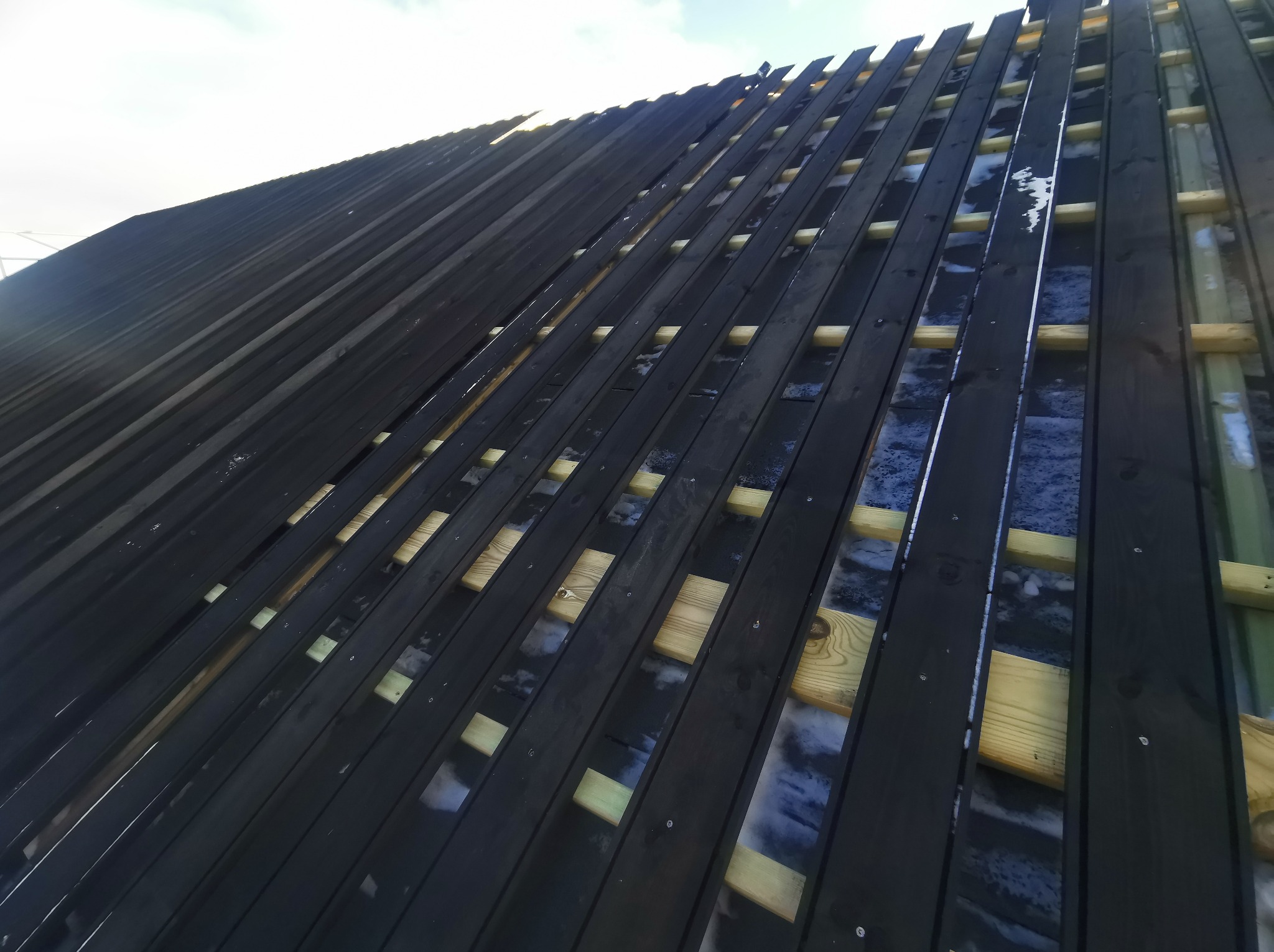 Konstrukcja dachu z czarnych desek i drewnianych łat, widoczna spod spodu z lekkim pochyleniem, ukazująca etap budowy.