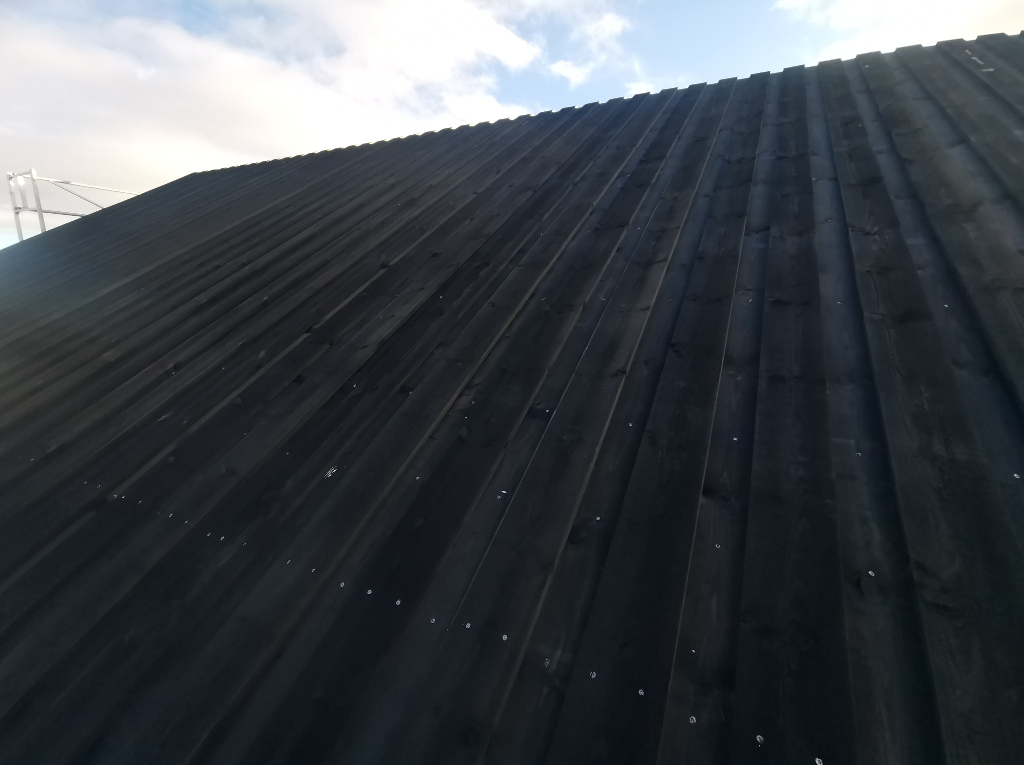 Ciemna, drewniana elewacja domu z widocznymi łączeniami desek i fragmentem rusztowania w tle, uchwycona pod kątem.