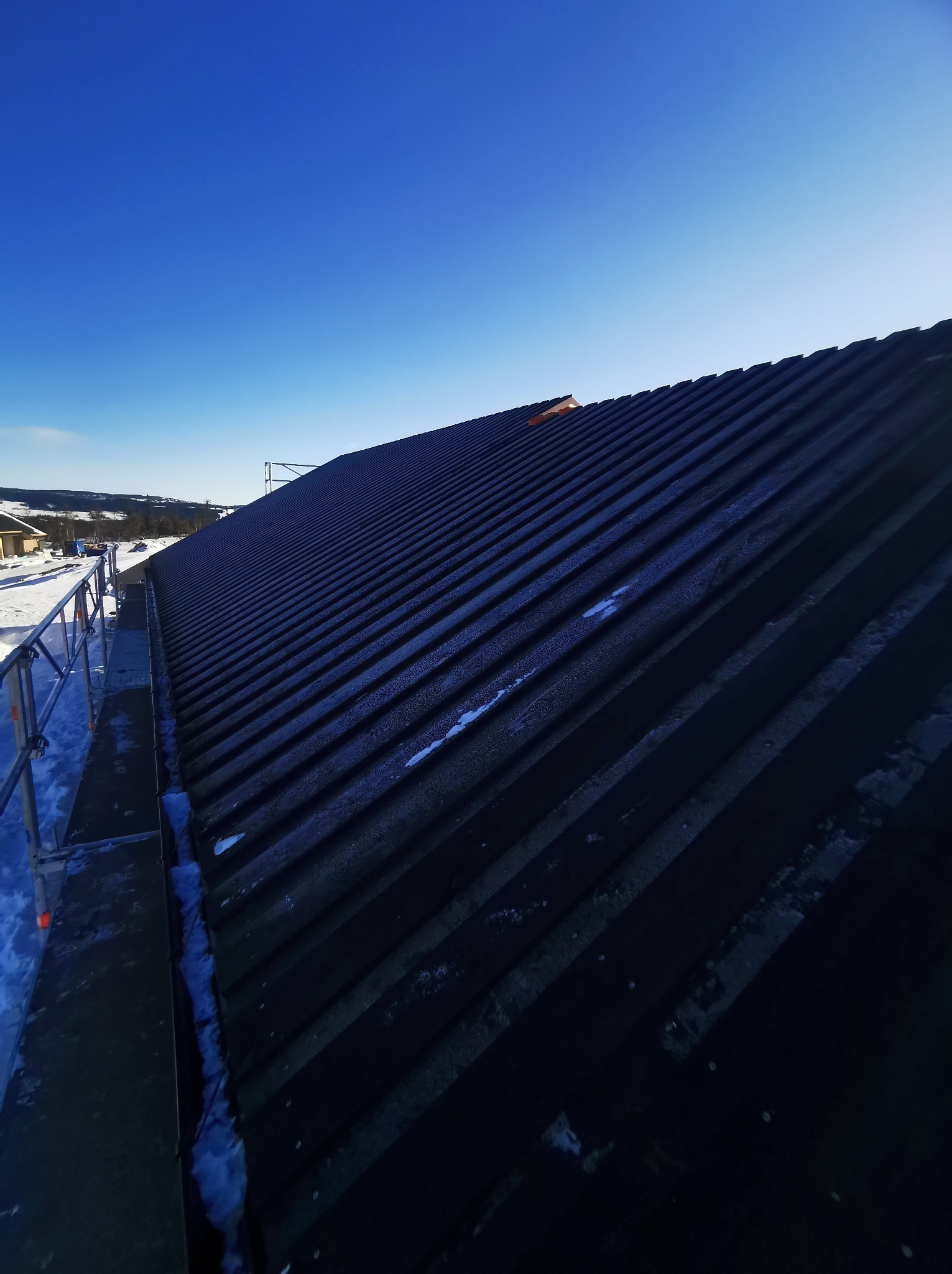 Ciemna, metalowa powierzchnia dachu z widocznym rusztowaniem i śniegiem w tle, na tle błękitnego nieba.