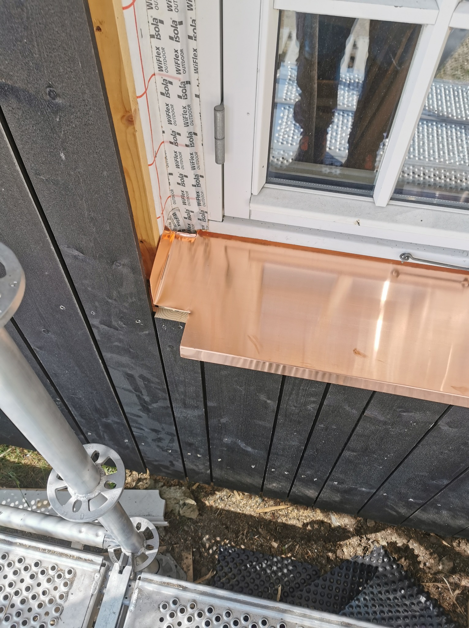 Montaż miedzianego parapetu zewnętrznego pod oknem na elewacji z ciemnego drewna, widoczne rusztowanie.