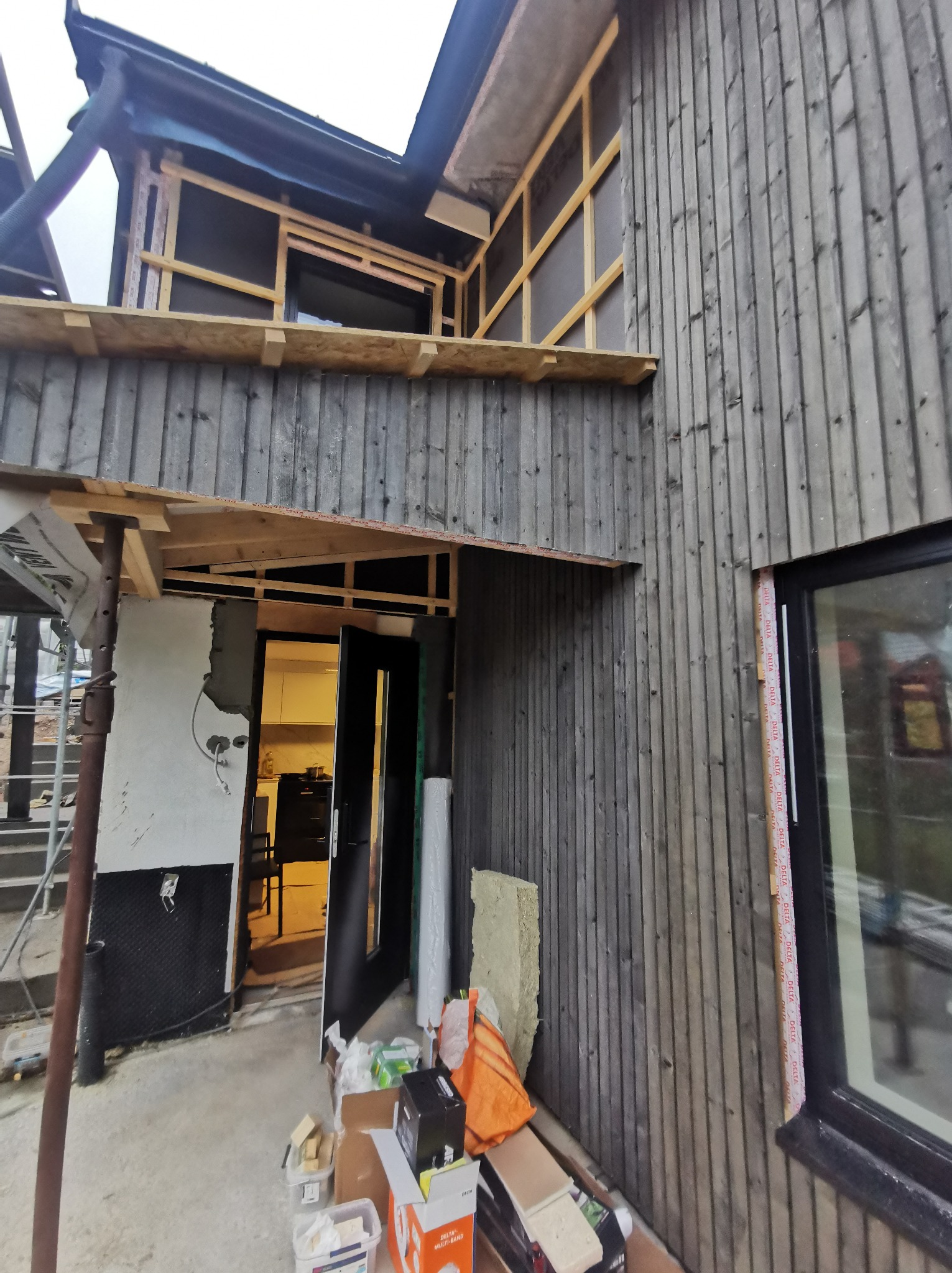 Budowa domu szkieletowego z elewacją z desek, widoczne rusztowania, materiały budowlane i otwarte drzwi do wnętrza z widoczną kuchnią.