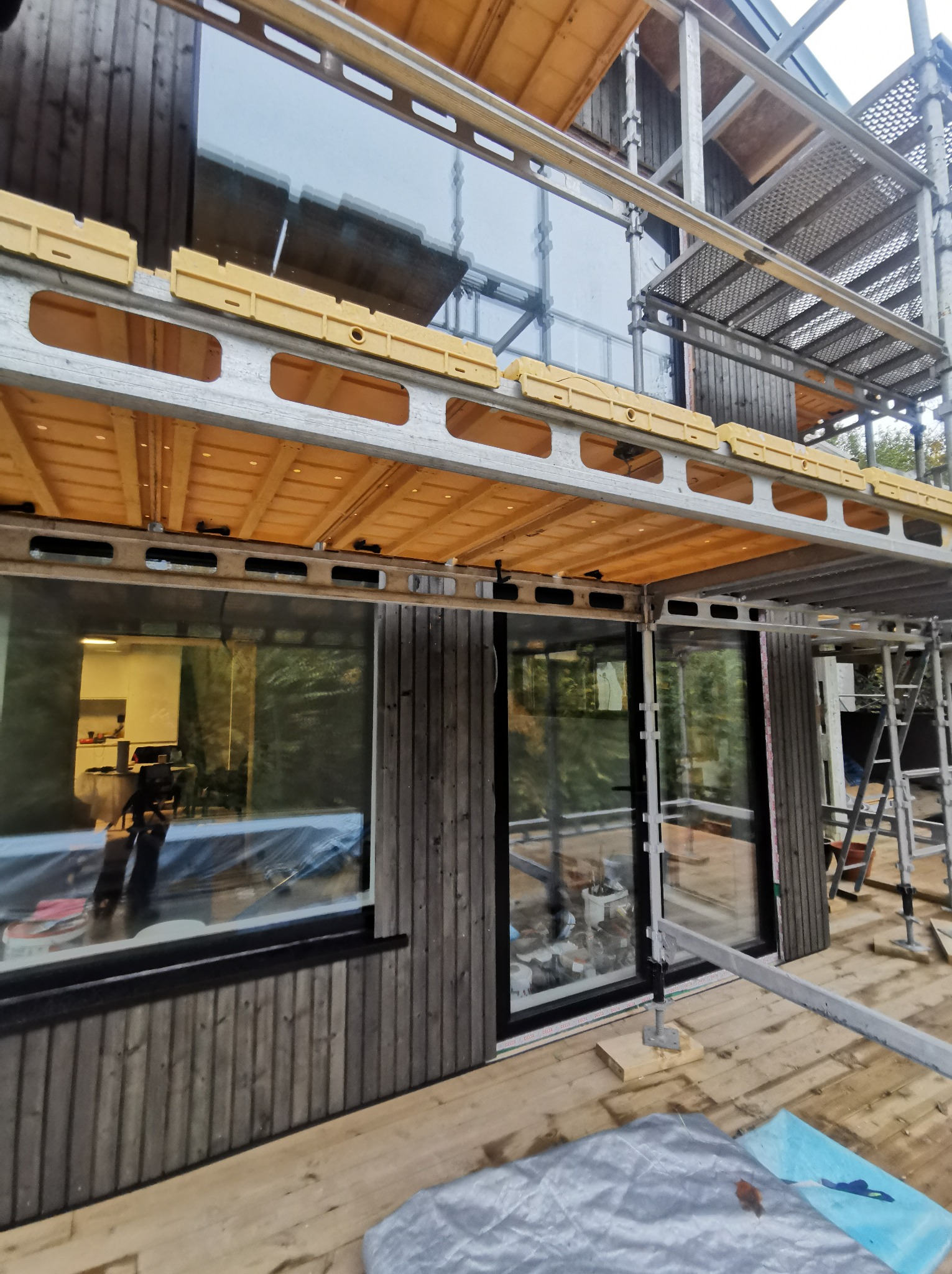 Budowa domu: fragment elewacji z szarego drewna, okna w czarnych ramach, widoczne rusztowania i materiały budowlane, wnętrze widoczne przez okno w trakcie wykańczania.